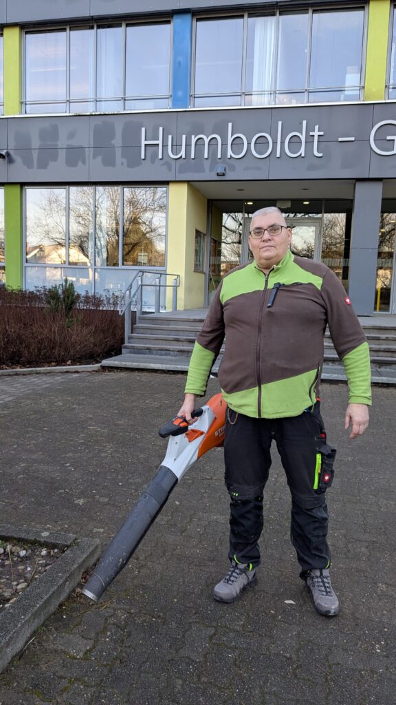 Foto des Schulhausmeisters, Herrn Böhm, der mit einem Laubsauger in der Hand vor dem Haupteingang des HGK steht.