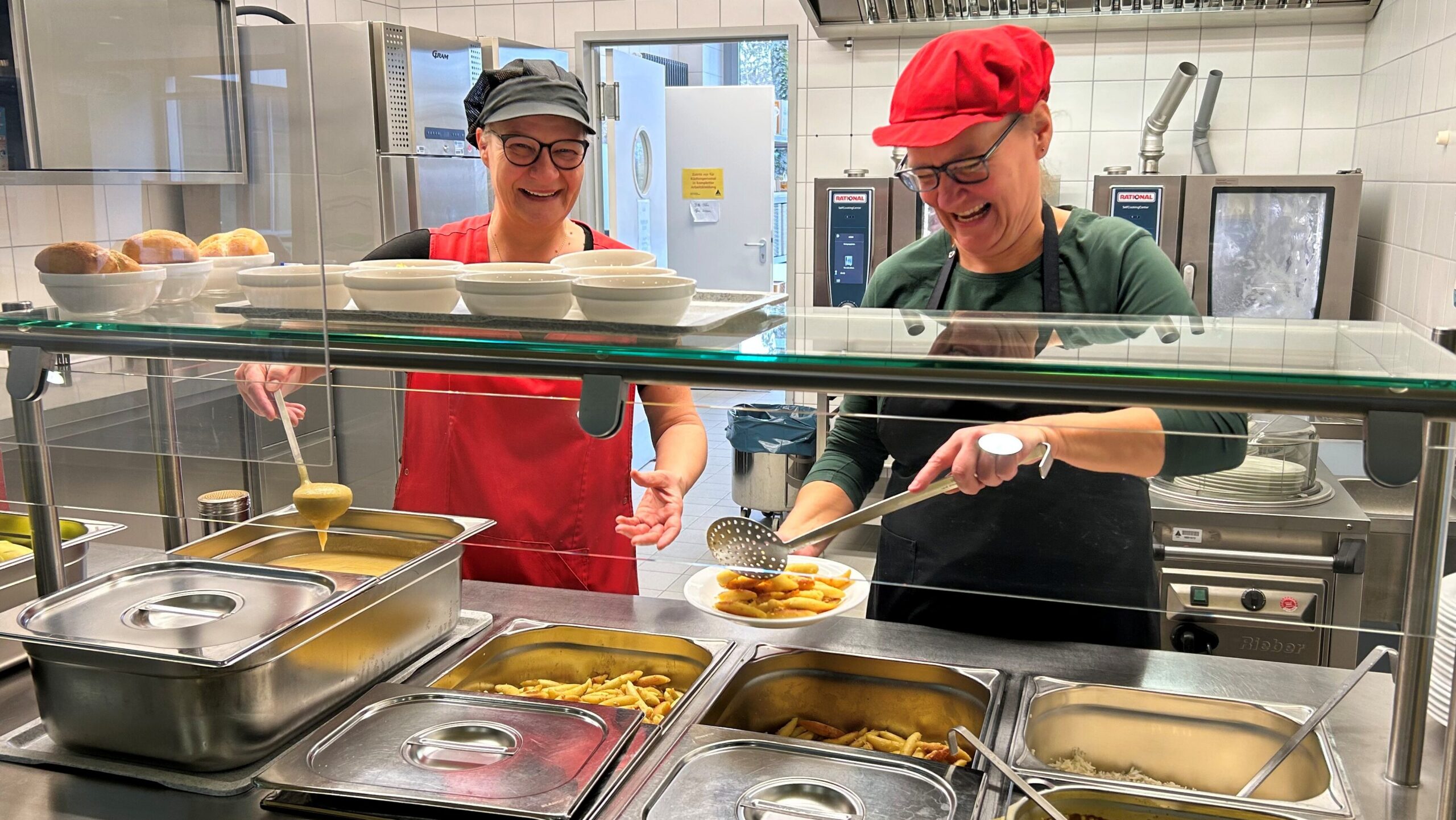 Die beiden Mensa-Köchinnen stehen gut gelaunt hinter ihrem Tresen und geben Mittagessen aus.