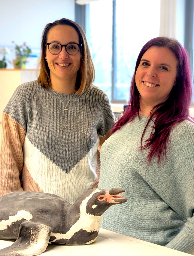 Foto der beiden Sekretärinnen, Frau Bergmann (links) und Frau Maile, die lächelnd hinter einem Pinguin-Modell auf dem Tresen vor ihnen stehen.